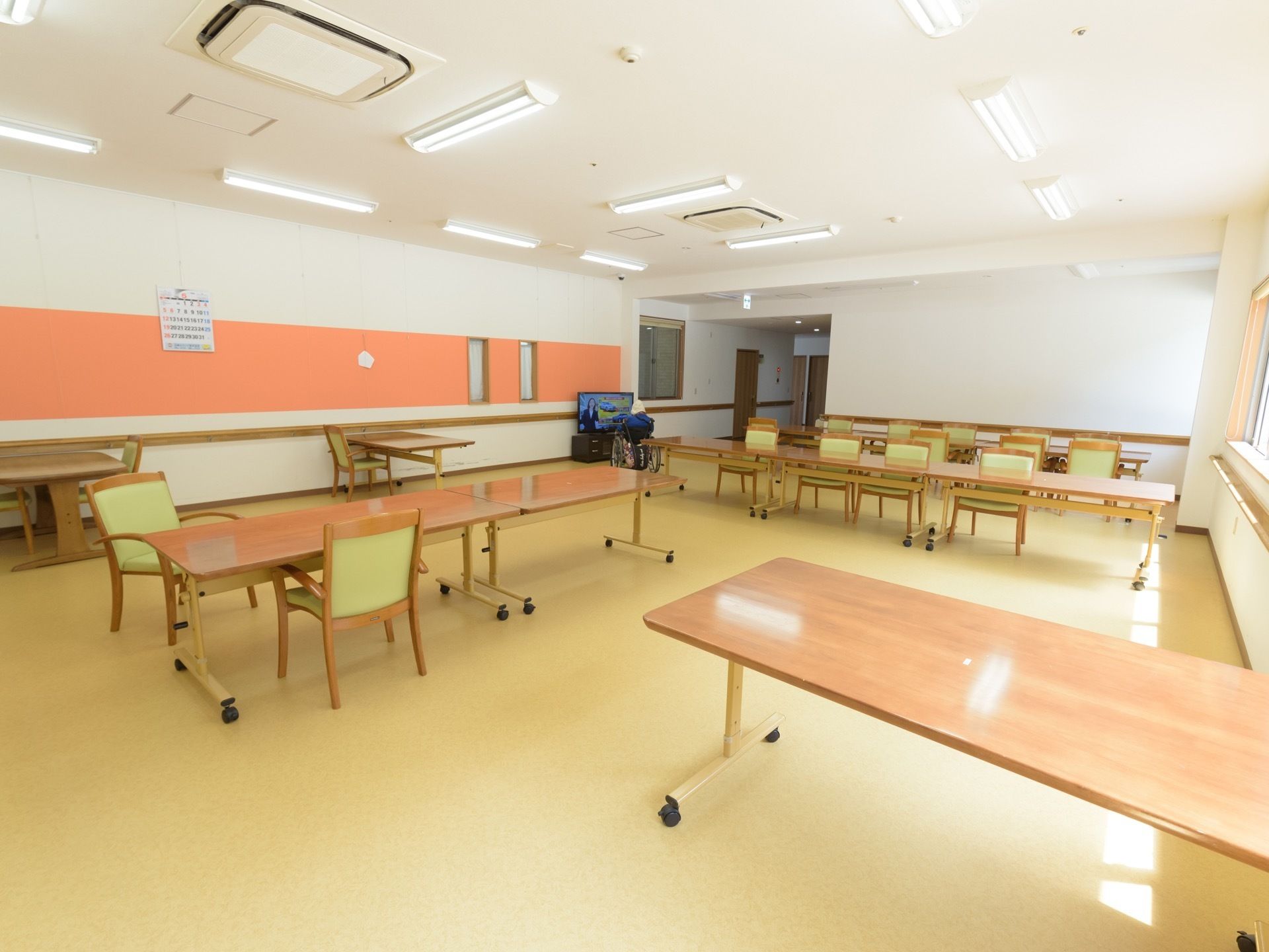 食堂に多くのテーブルと椅子が並べられている。壁には月間カレンダーが掛けられている。テレビが設置されている。