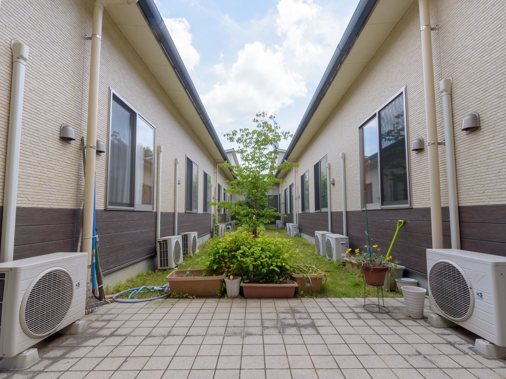 建物に囲まれた中庭がある。芝生の中央部に背の高い木が植えられている。エアコンの室外機が多く置かれている。