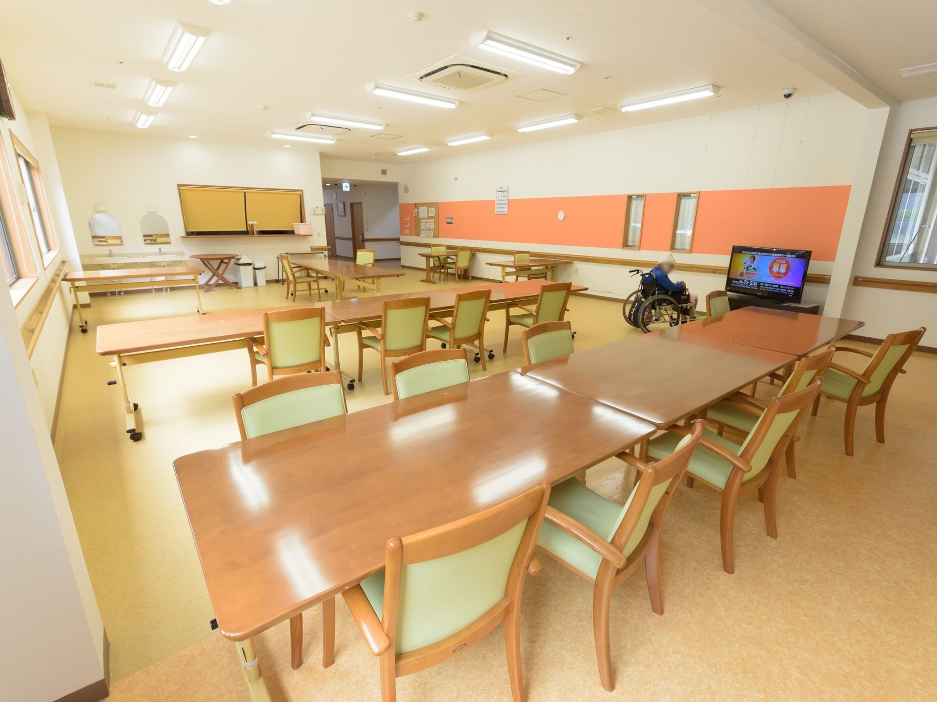 食堂に多くの椅子とテーブルが置かれている。壁際に設置されたテレビの前に、車椅子をご利用の入居者様がお一人おられる。