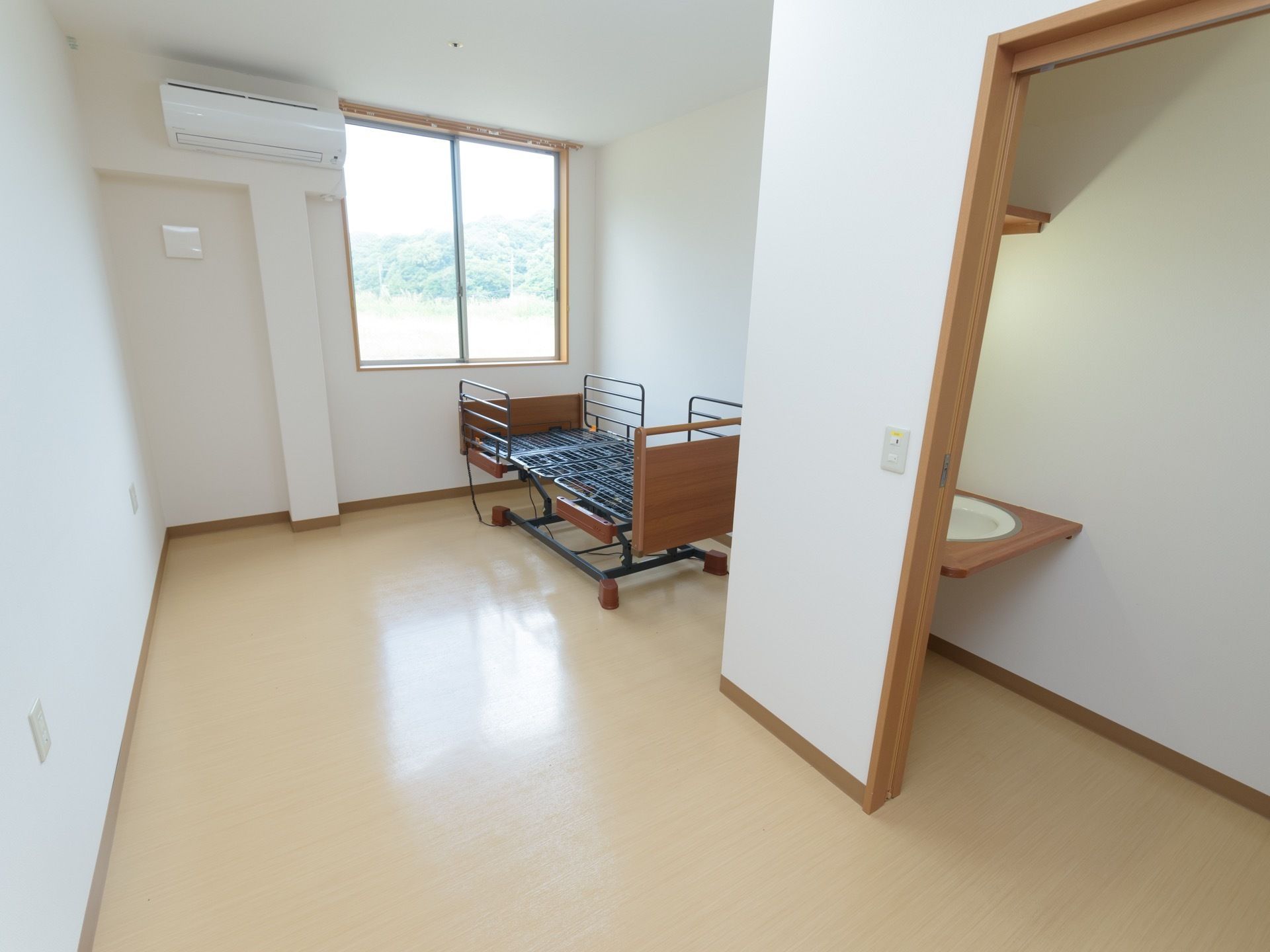 腰高窓の下に手すりがついた介護ベッドが置いてある。真っ白な壁にエアコンと換気扇が設置されている。扉の奥に洗面台が見える。
