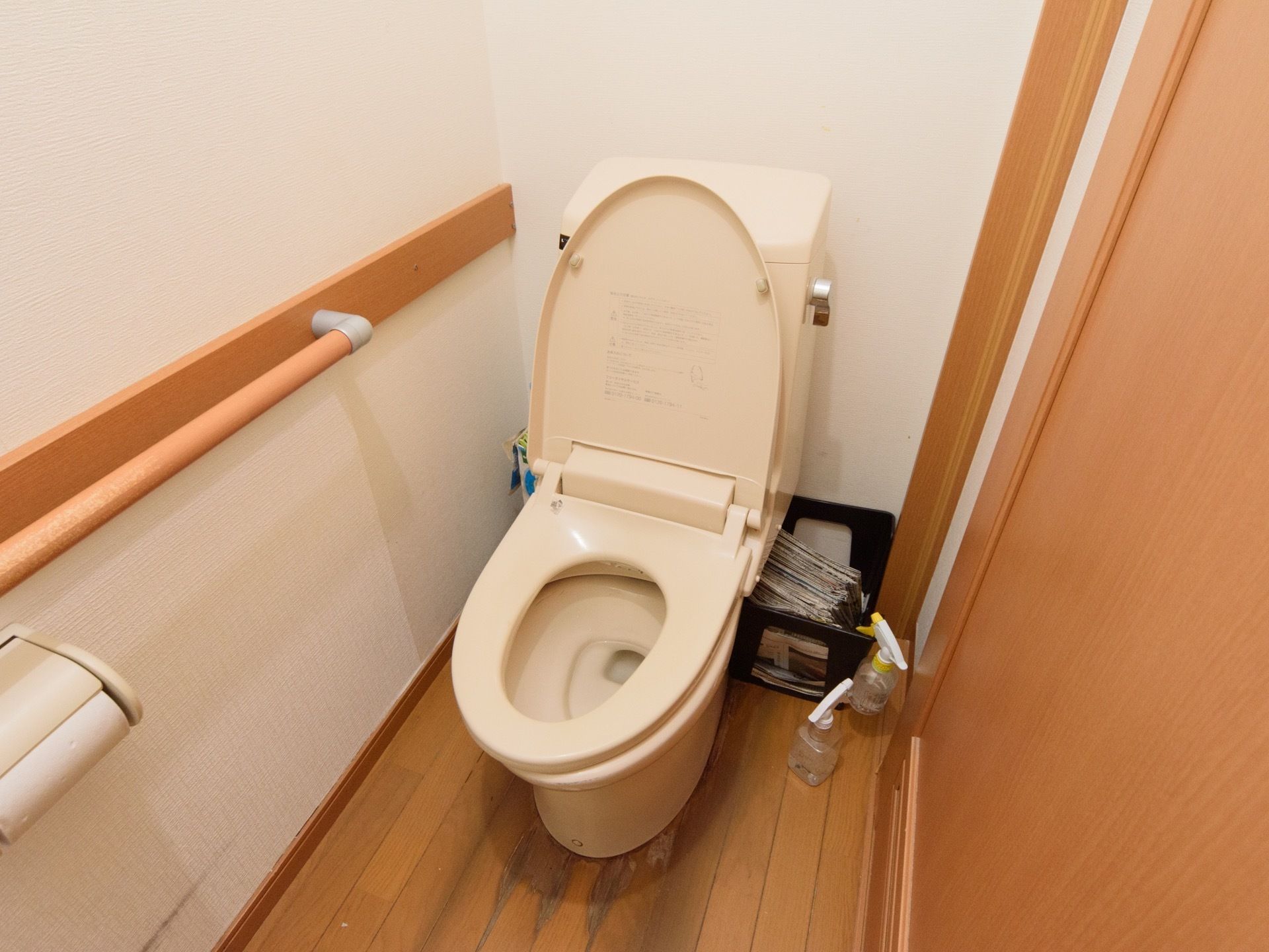 白壁にフローリングの内装をもつ個室トイレである。便座横の壁には、しっかりとした手すりが設置されている。