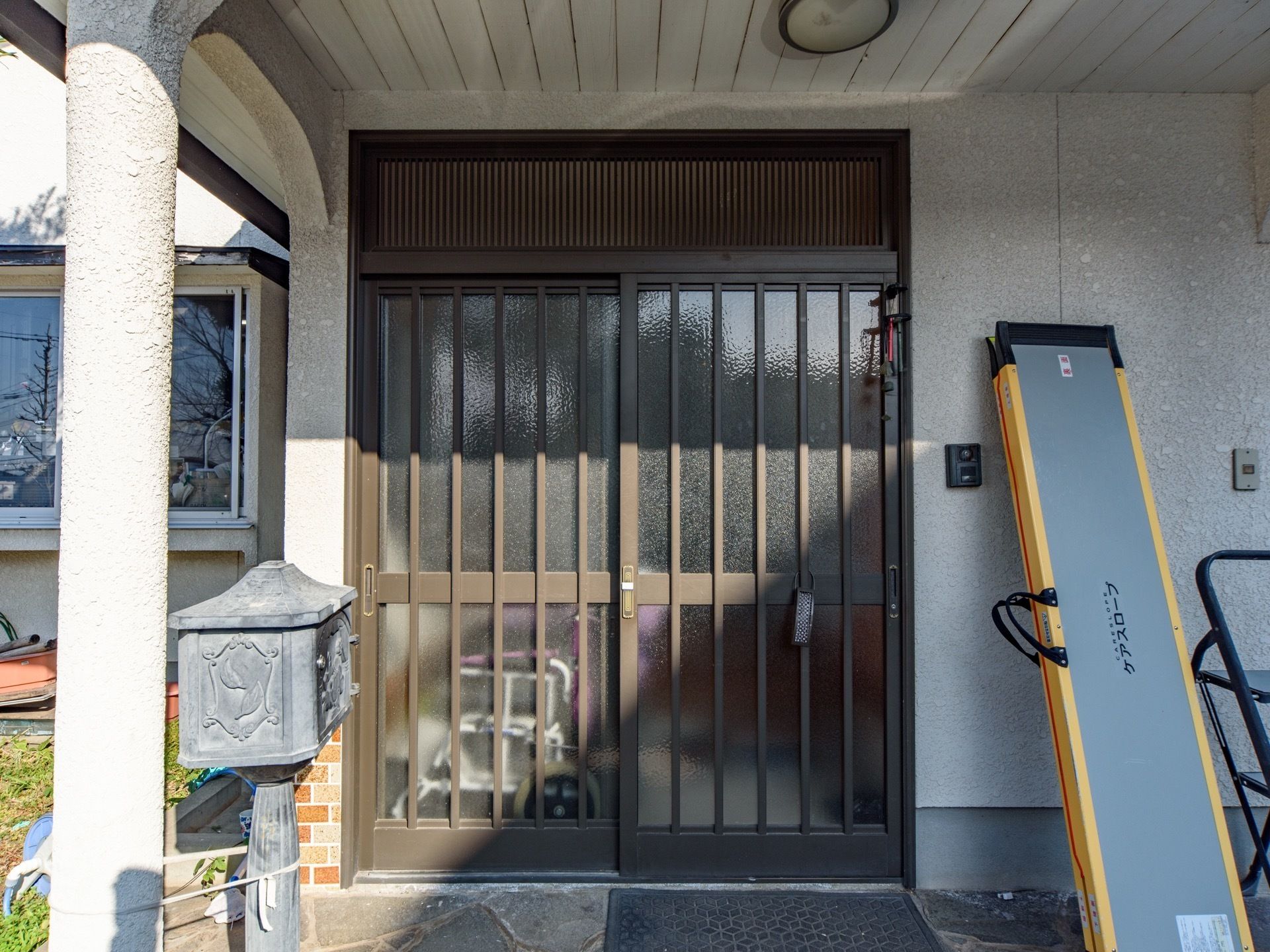 引き違い戸が付いた玄関の前に、マットが敷いてある。カメラがあり、来訪者様を確認できるインターフォンも設置している。