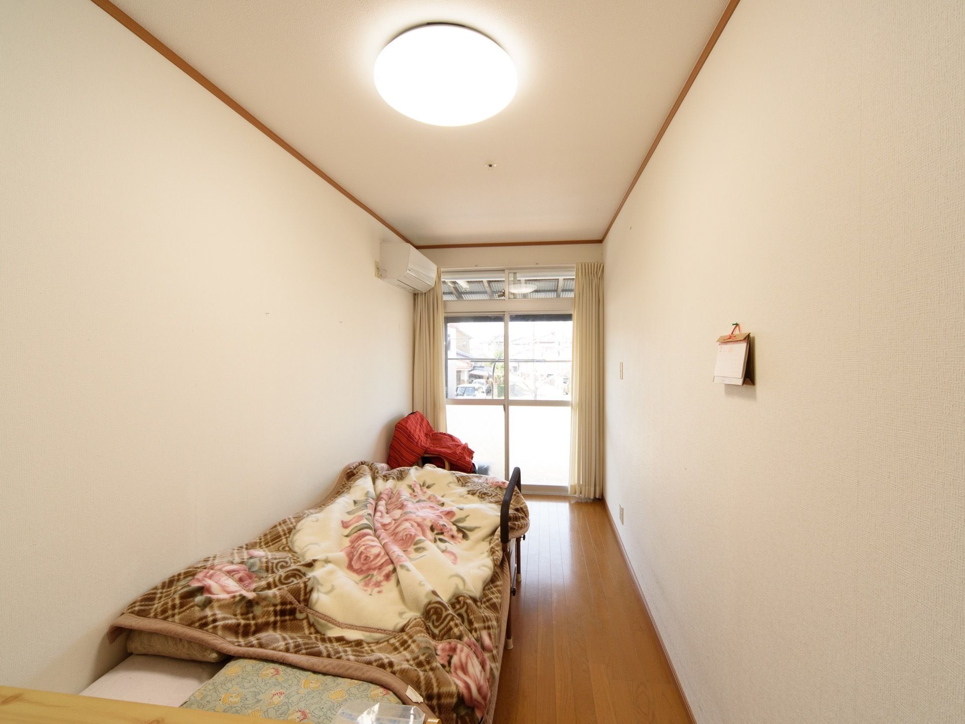 居室の奥は掃き出し窓になっており、クリーム色のカーテンが掛けられている。その隣にはエアコンが設置されている。