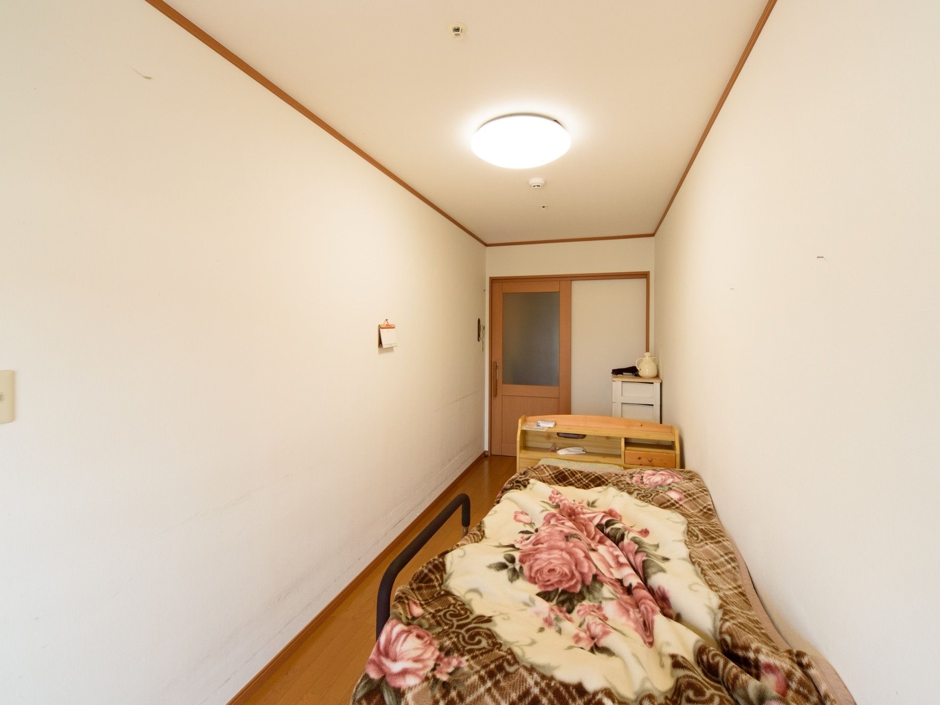 居室は縦長の間取りであり、チェストとベッドが置かれている。木製のヘッドボードには棚があり、備品を置くことができる。
