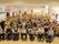 大勢の高齢者がフローリングの共有スペースに集まっている。椅子に座っている人もいれば、車いすの人もいる。