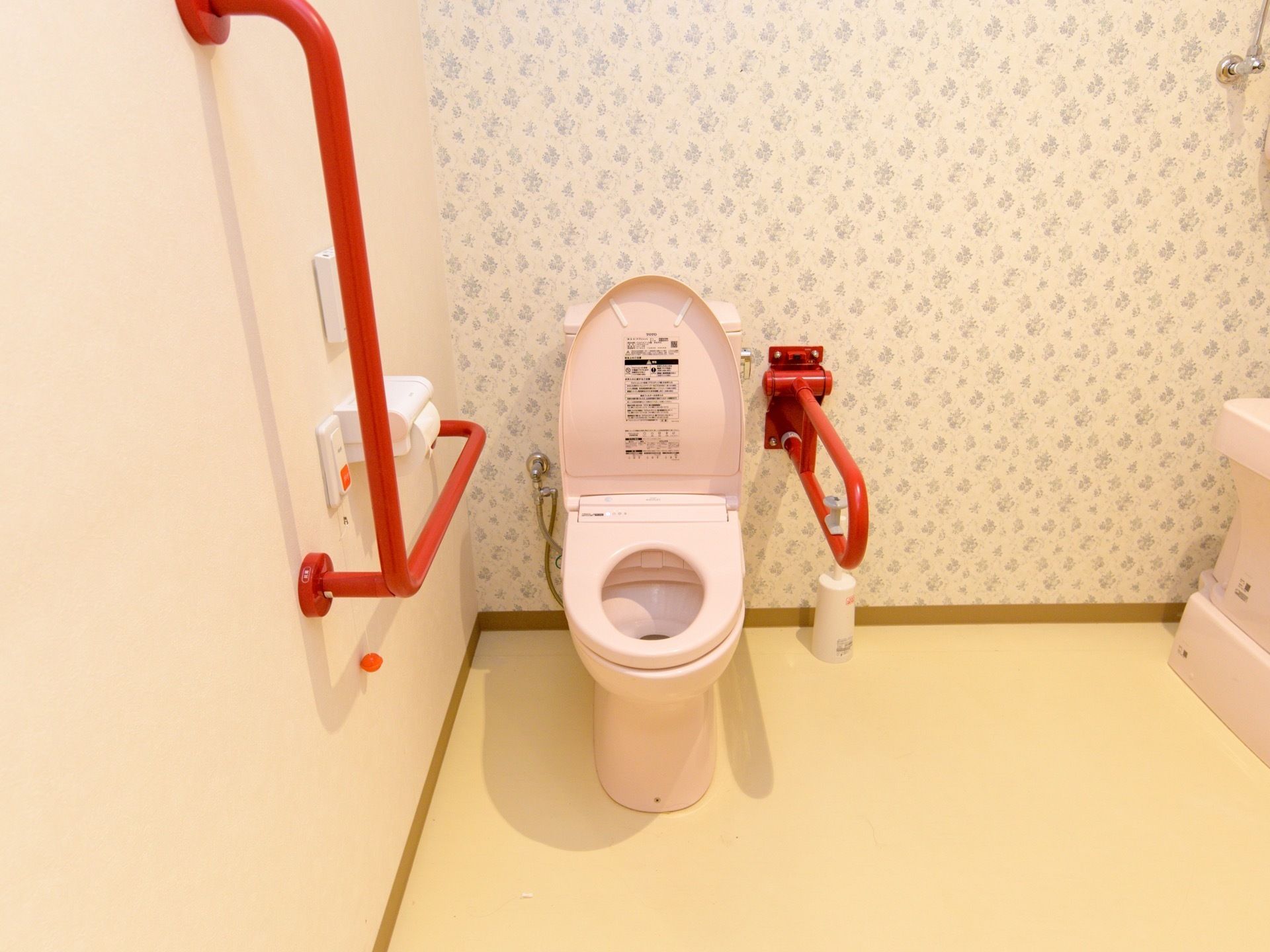 トイレはピンク色の便座になっている。鮮やかな赤色の手すりが、壁や便座脇に設置されている。緊急呼び出しボタンがある。
