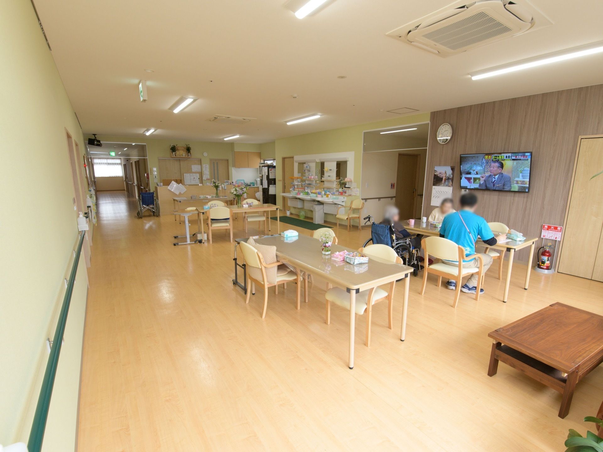 食堂には、多くのテーブルと椅子が置かれている。女性の入居者様２名と、男性スタッフ１名が同じテーブルに座っている。