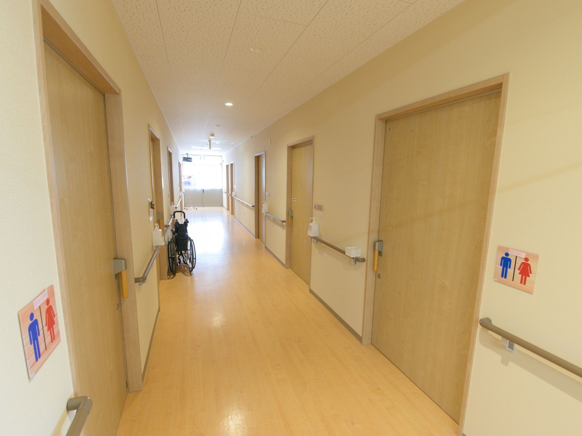 廊下は、つきあたりの窓からの採光で明るくなっている。居室入口付近の手すりの上に、消毒用アルコールのボトルが置かれている。