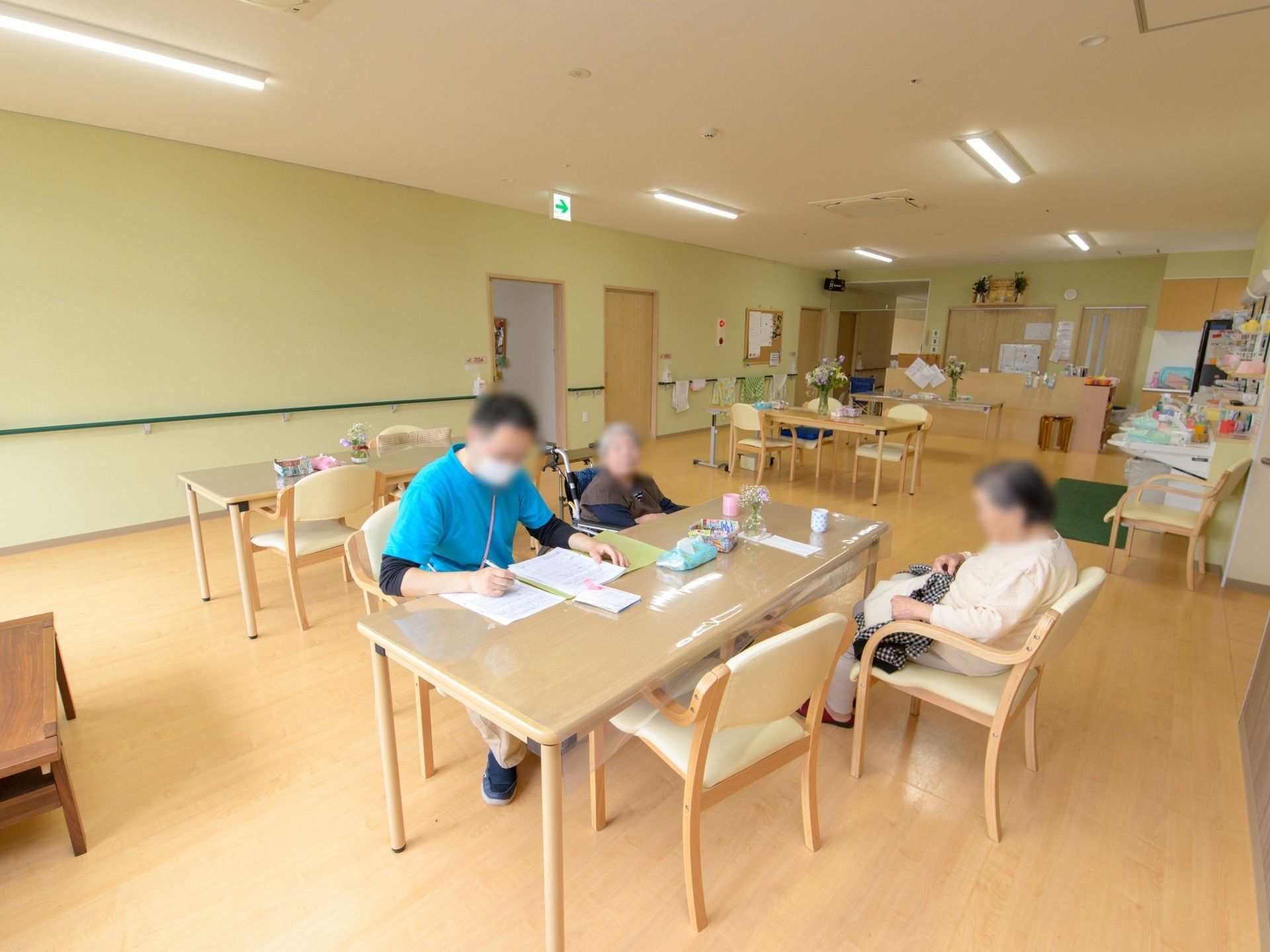 食堂の片隅のテーブルに、入居者様２名とスタッフ１名が集まっている。入居者様のお一人は、車椅子のままでテーブルについている。