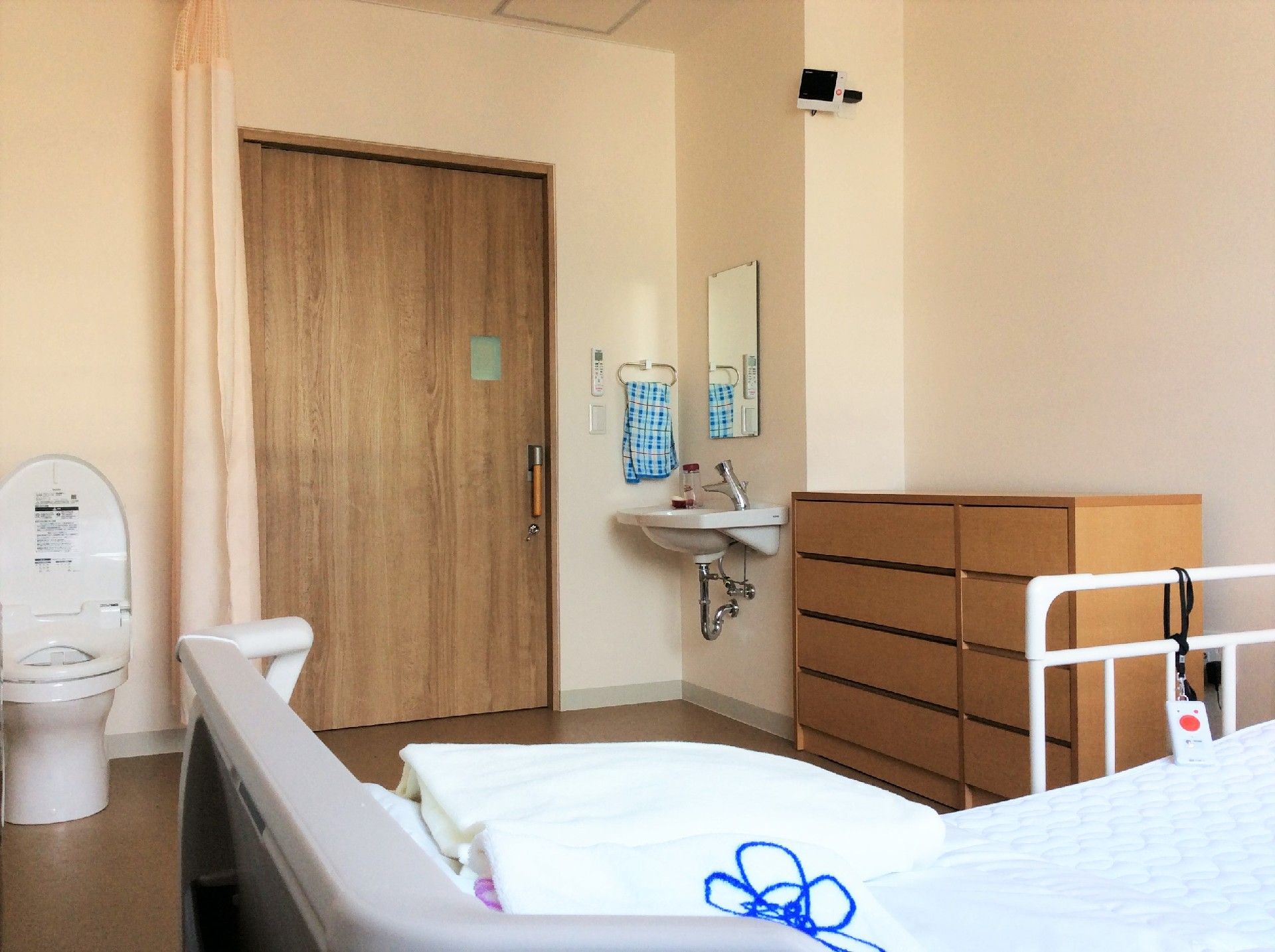 居室は木目調のドアに薄いピンクの壁紙が柔らかい雰囲気。トイレや洗面所も付き、手前にはベッドやタンスが置かれている。