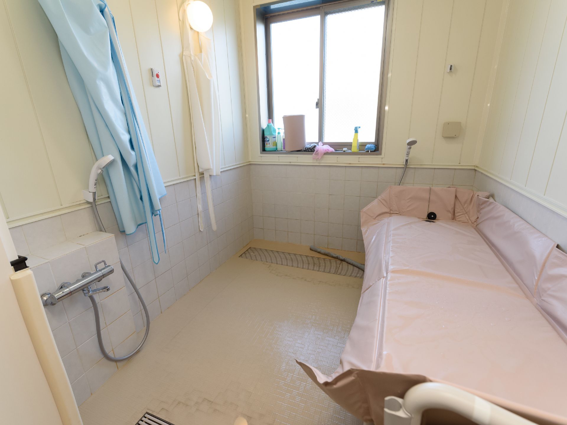 洗い場にシャワーが設置されている。壁にはスタッフ用の介助エプロンがかかっている。窓から柔らかな光が差し込んでいる。