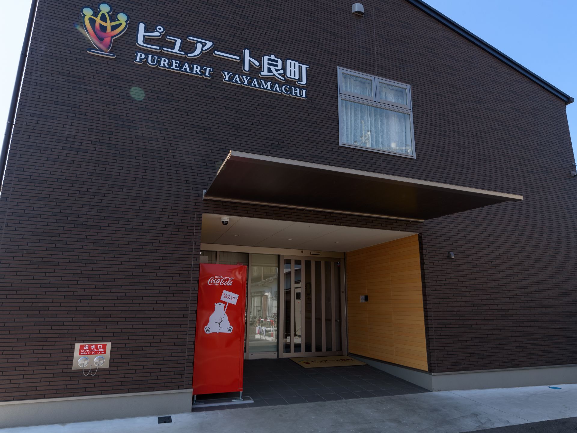 濃い茶系の外壁の建物で、玄関上部の壁に施設名が大きく表示されている。玄関には屋根があり、自動販売機が置かれている。