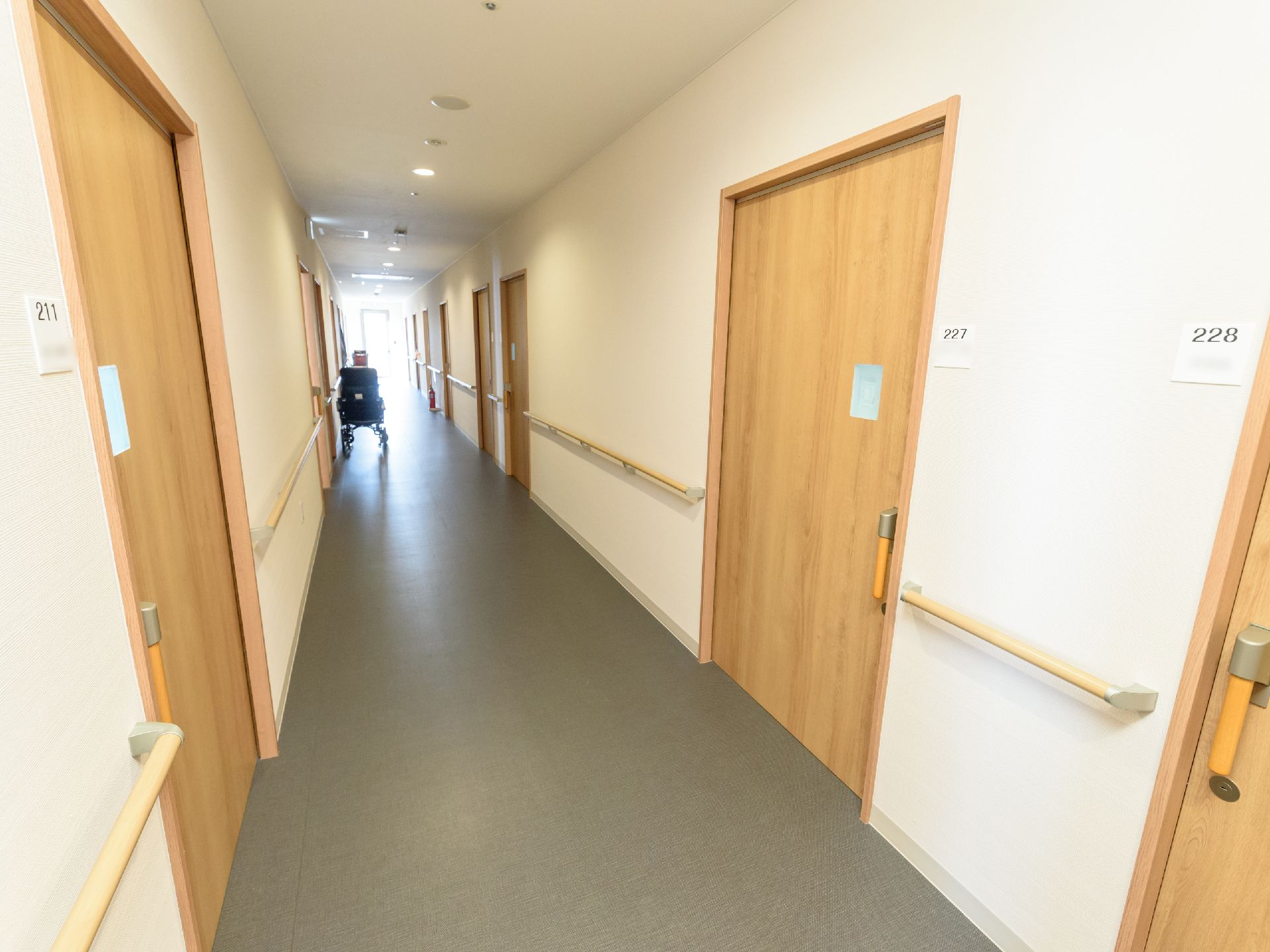 廊下の両側の壁に、居室のドアと手すりが並んでいる。ドアは引き戸で、横の壁に部屋番号が表示されている。