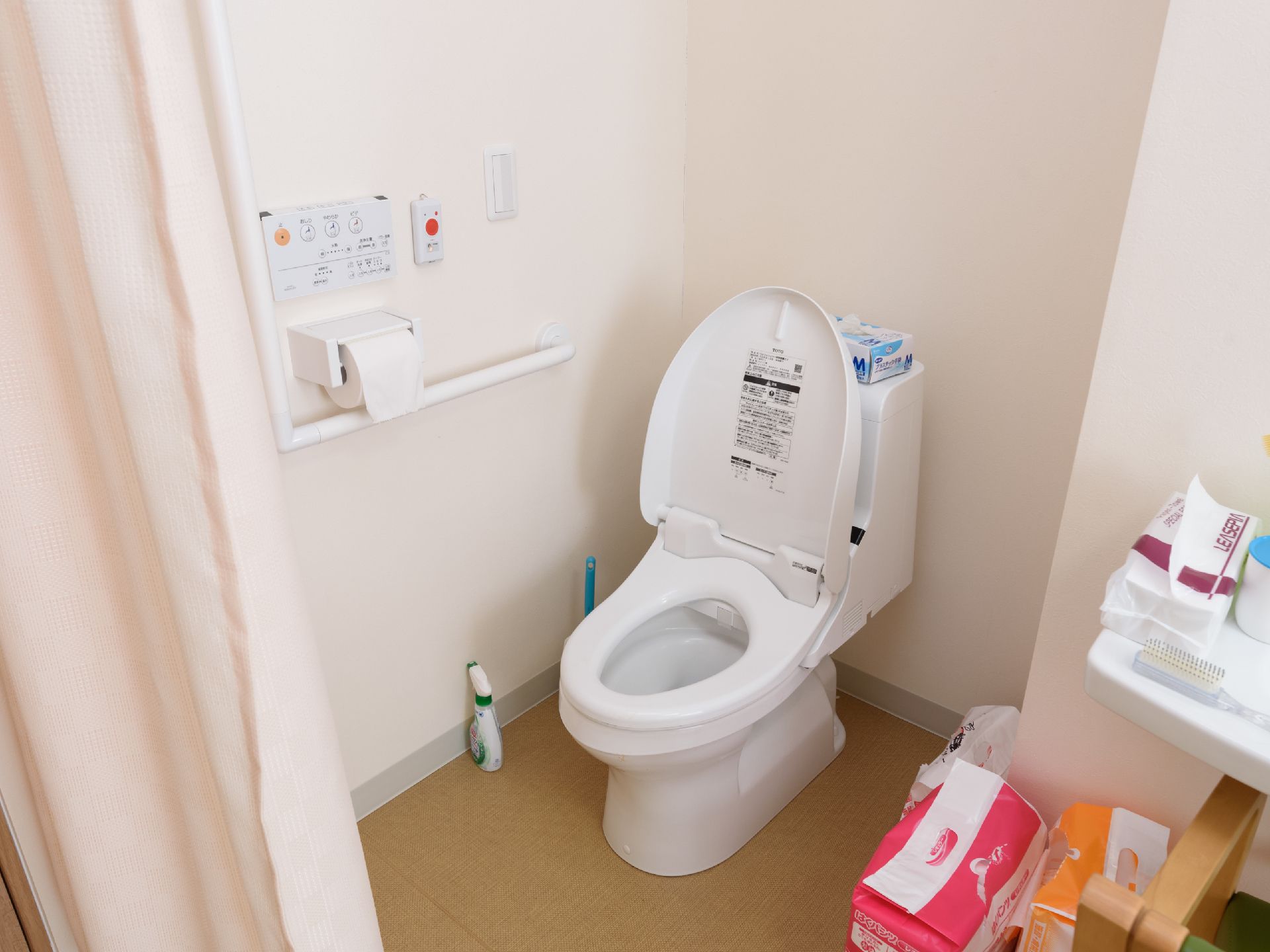 温水洗浄機付きの洋式トイレで、壁には手すりや緊急コールが設置されている。個室はカーテンで仕切られている。