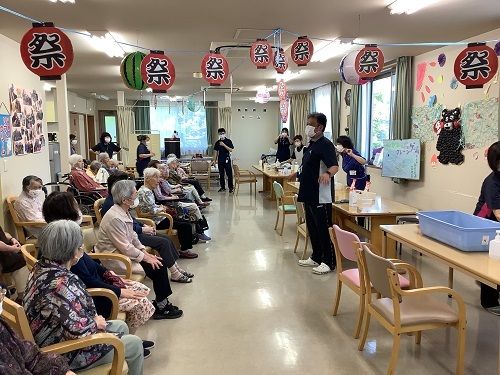 テーブルが複数用意され、それぞれにスタッフがいる。イベントの前にスタッフの話を聞いている入居者たち。