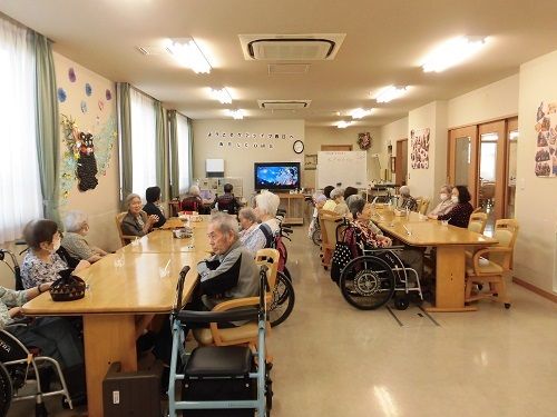 複数のテーブルが並べられ、２列の大きなテーブルとなっている。それぞれのテーブルにたくさんの高齢者がいる。