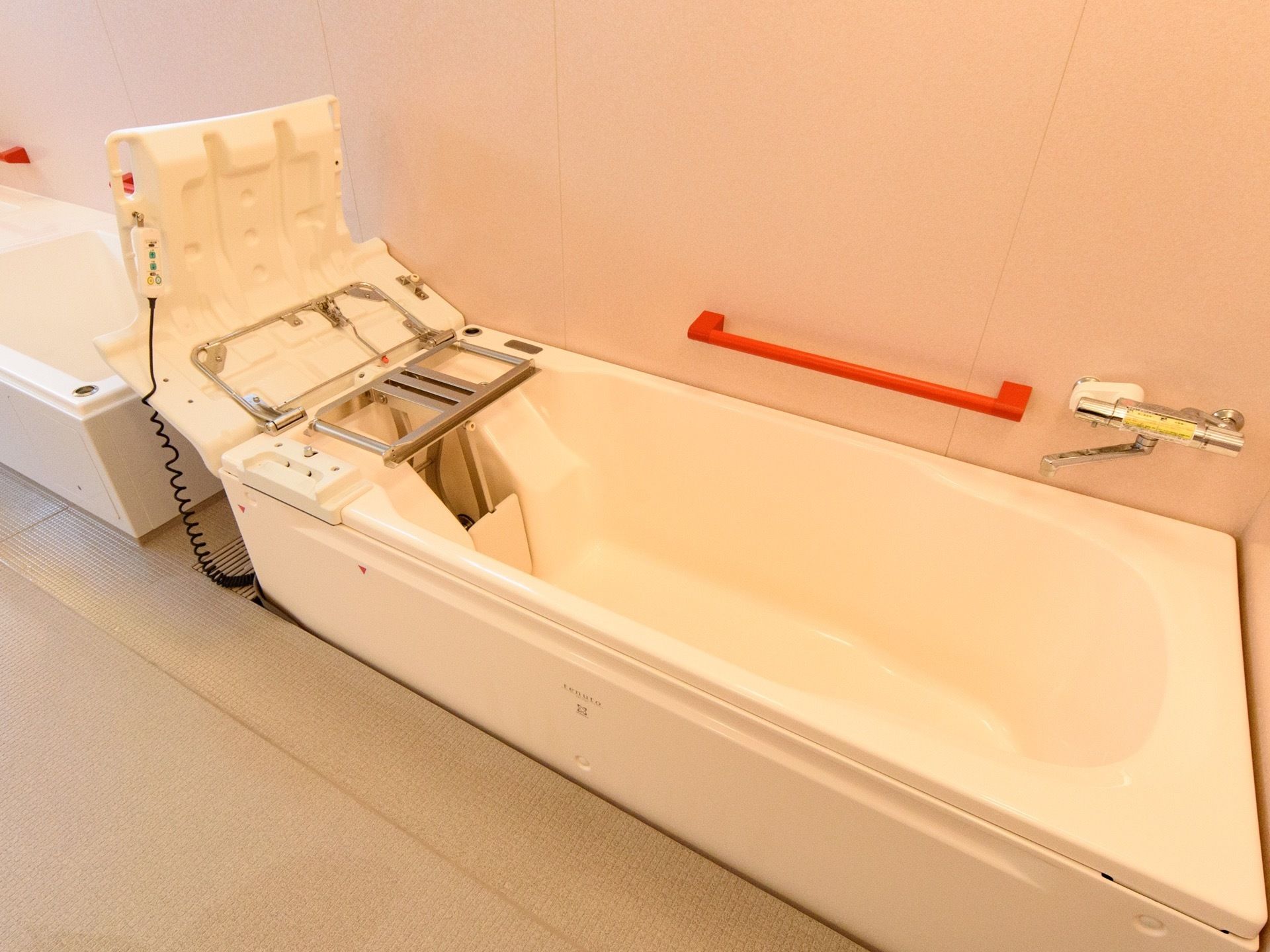 ベージュを基調とした室内に、入浴用の補助具の付いた長い浴槽が置かれている。浴槽脇にはオレンジ色の手すりがある。