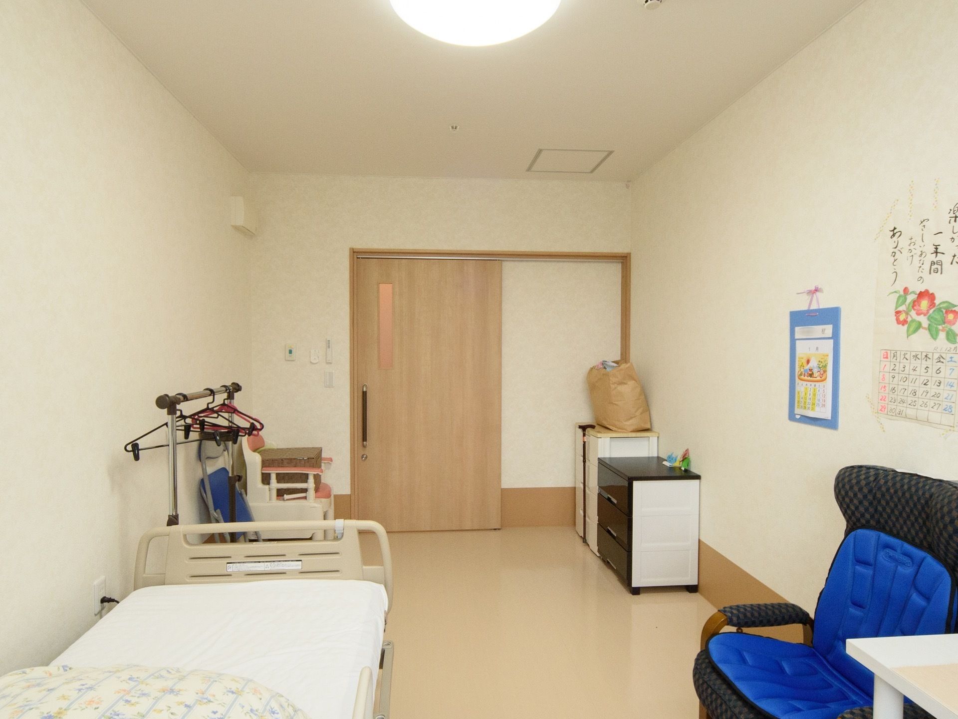 ベージュが基調の室内に、ライトブラウンの引き戸が付いている。ベッドと青い１人掛けのソファが置かれている。