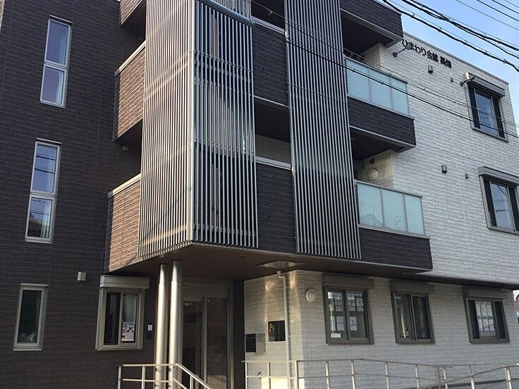 モノトーンの3階建ての建物の玄関には手すり付きのスロープがあり、各フロアにはベランダも設置されている。