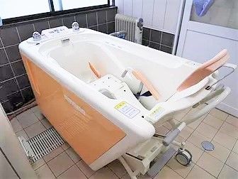 白とピンクの機械浴室が設置された浴室は黒とブラウンのタイル張り。介護度が高い方も入浴できる設備。