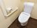 ダークブラウンのフローリングに、薄いクリーム色の壁のトイレである。壁にはコンセントと手すり、トイレットペーパーがある。