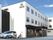 白を基調とした3階建ての建物の横には駐車場があり車が止まっていて、車いすの高齢女性がスタッフといる。