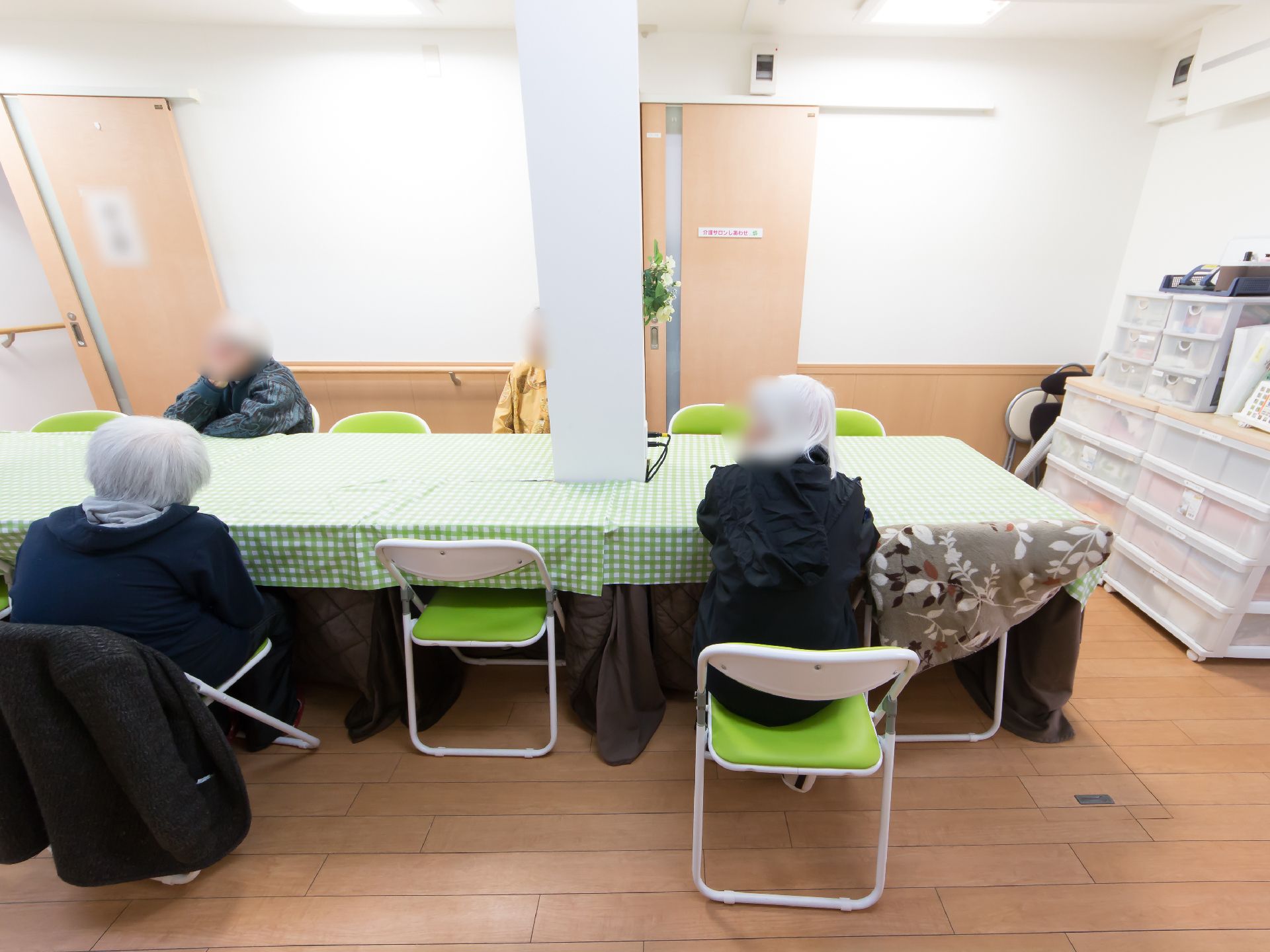 数人の入居者様がテーブルを囲んでいる。机の上にはテーブルクロスが敷かれており、横には収納ケースが置かれている。