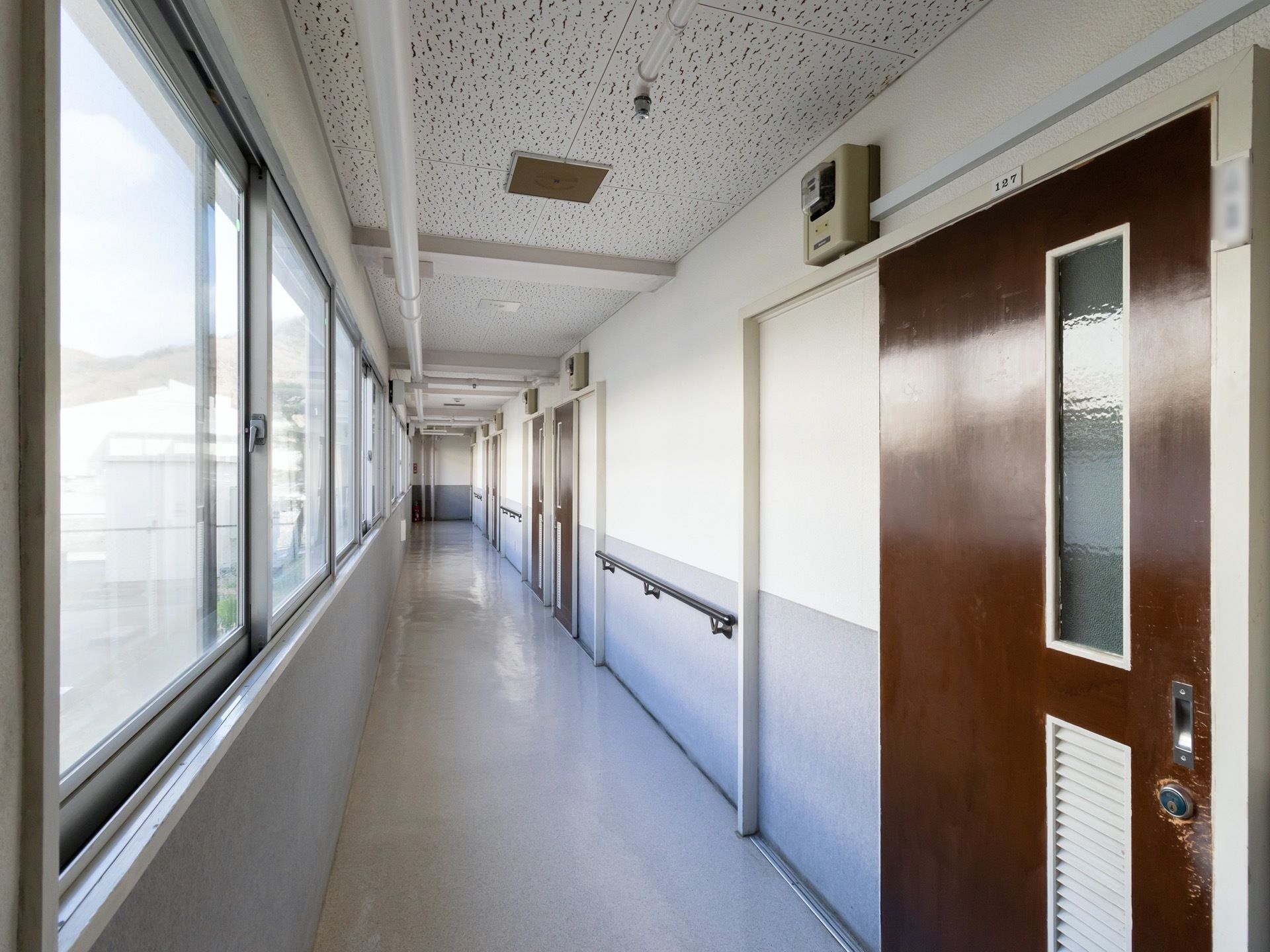 窓が片側にある明るい廊下である。もう片方には、居室の茶色い扉が並んでおり、扉の間の壁には手すりが備わっている。
