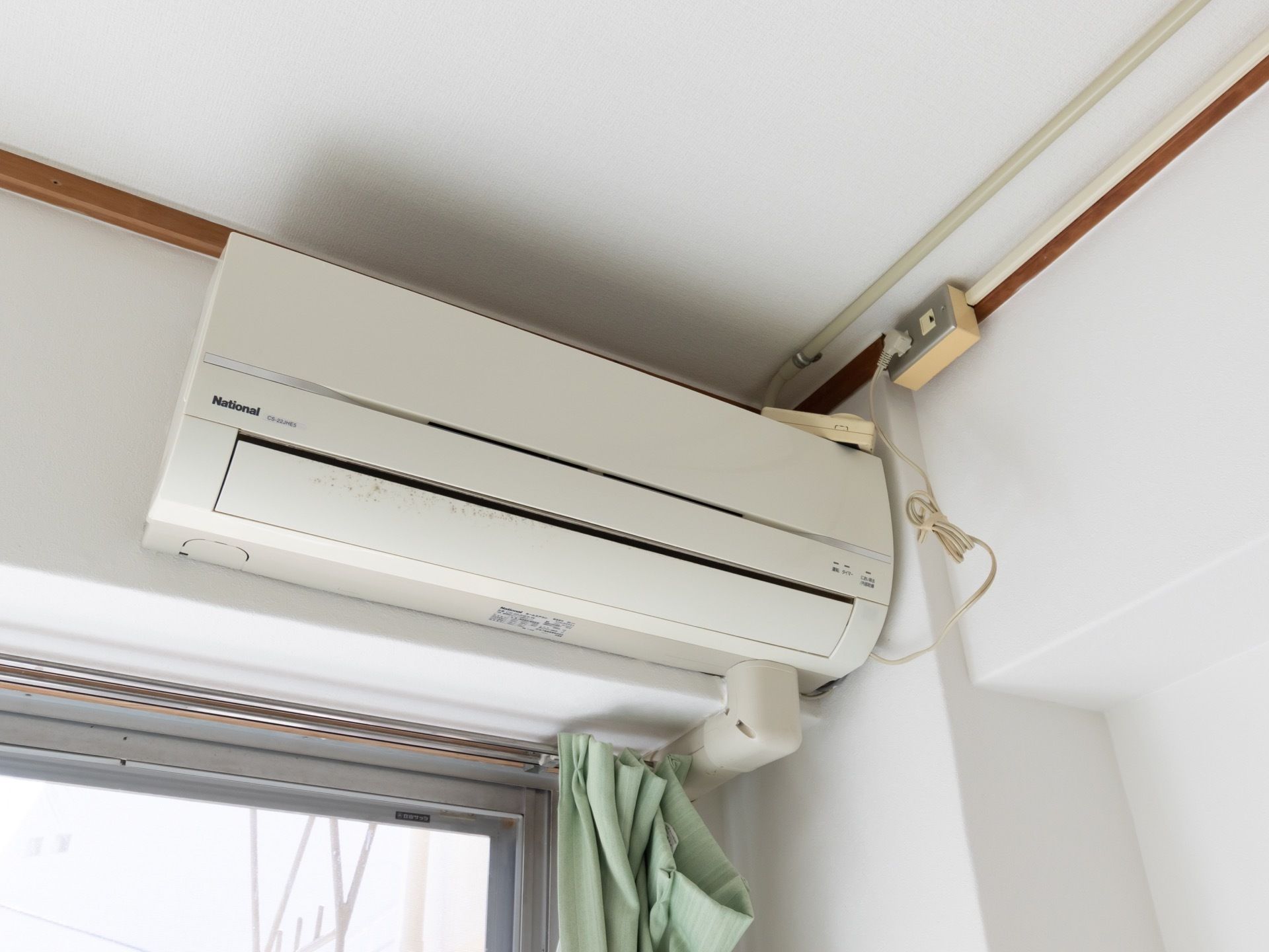 窓には緑色のカーテンが下がっており、その上に家庭用のエアコンが備えられている。窓の外には、物干し器具が設置されている。