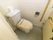 壁と床共にタイルが貼られたトイレがある。トイレは洋式タイプで、壁にトイレットペーパーフォルダーと手すりが備え付けられている。