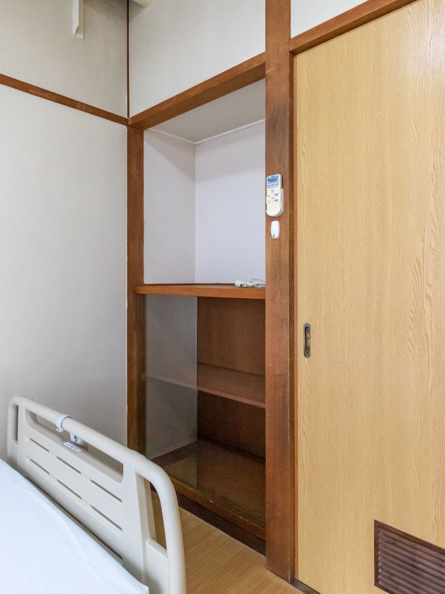 作り付けの棚が押し入れの隣にある。下側のみガラスのスライドドアがついている。柱にはエアコンのリモコンが収められている。