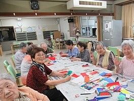 大きなテーブルを囲んで折り紙を使った工作をしている高齢者たち。カラフルな折り紙がテーブルの上にある。