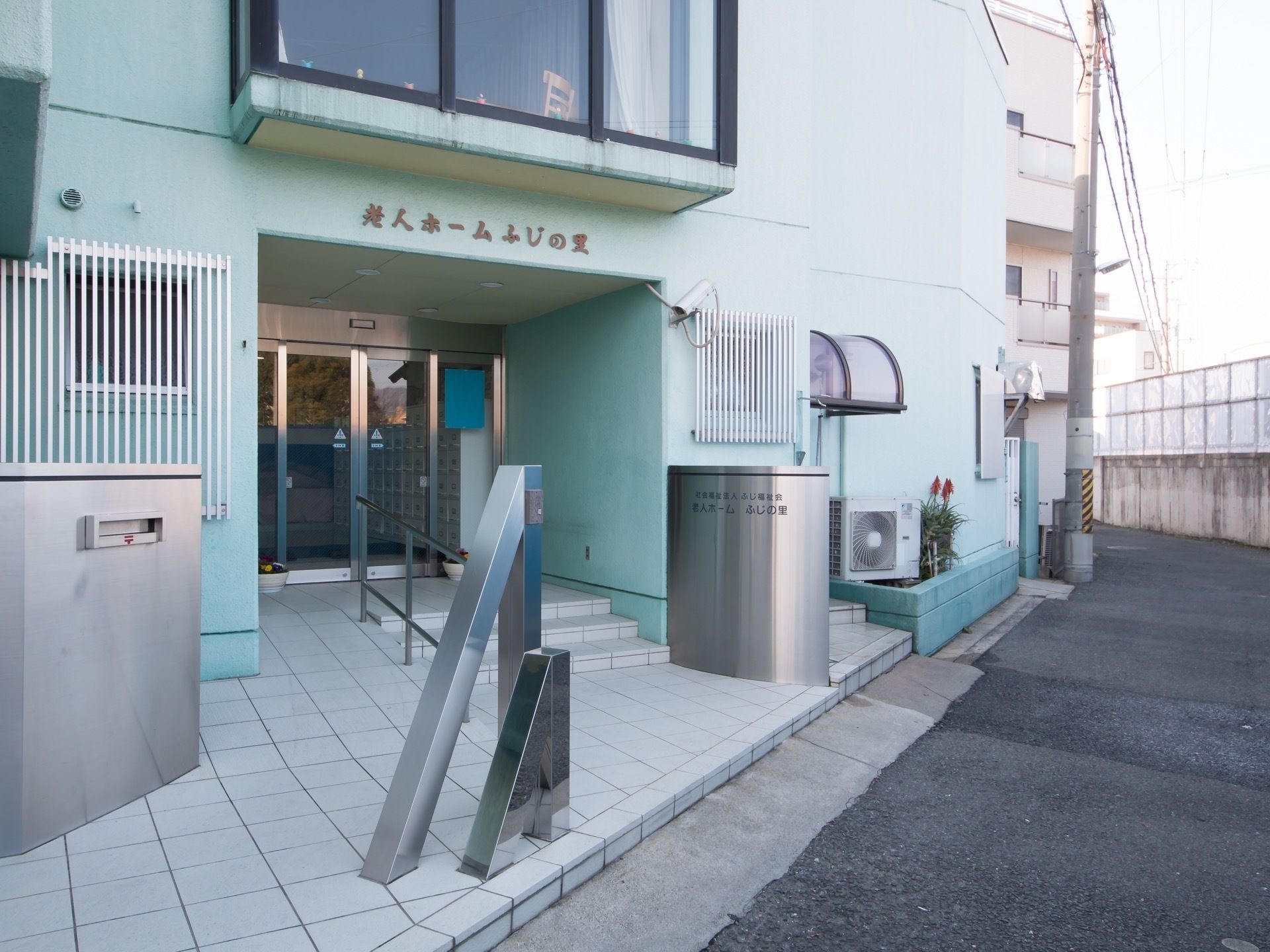 水色の外壁の建物が見える。建物の前方右側には看板とオブジェが設置されている。反対側には郵便受けが設けられている。