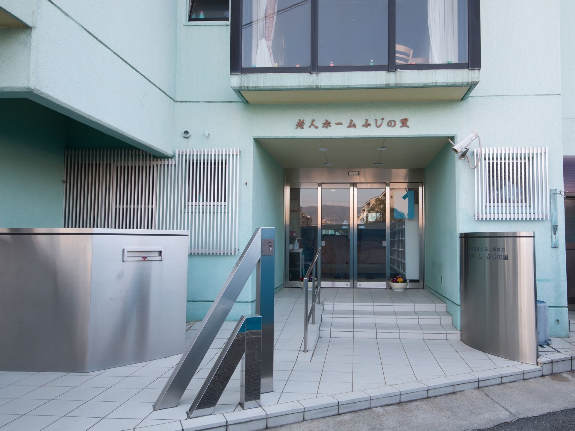 出入口は建物の少し奥まったところにあり、上には施設名が書かれている。玄関の前は白いタイル張りになっており、階段とスロープが設置されている。
