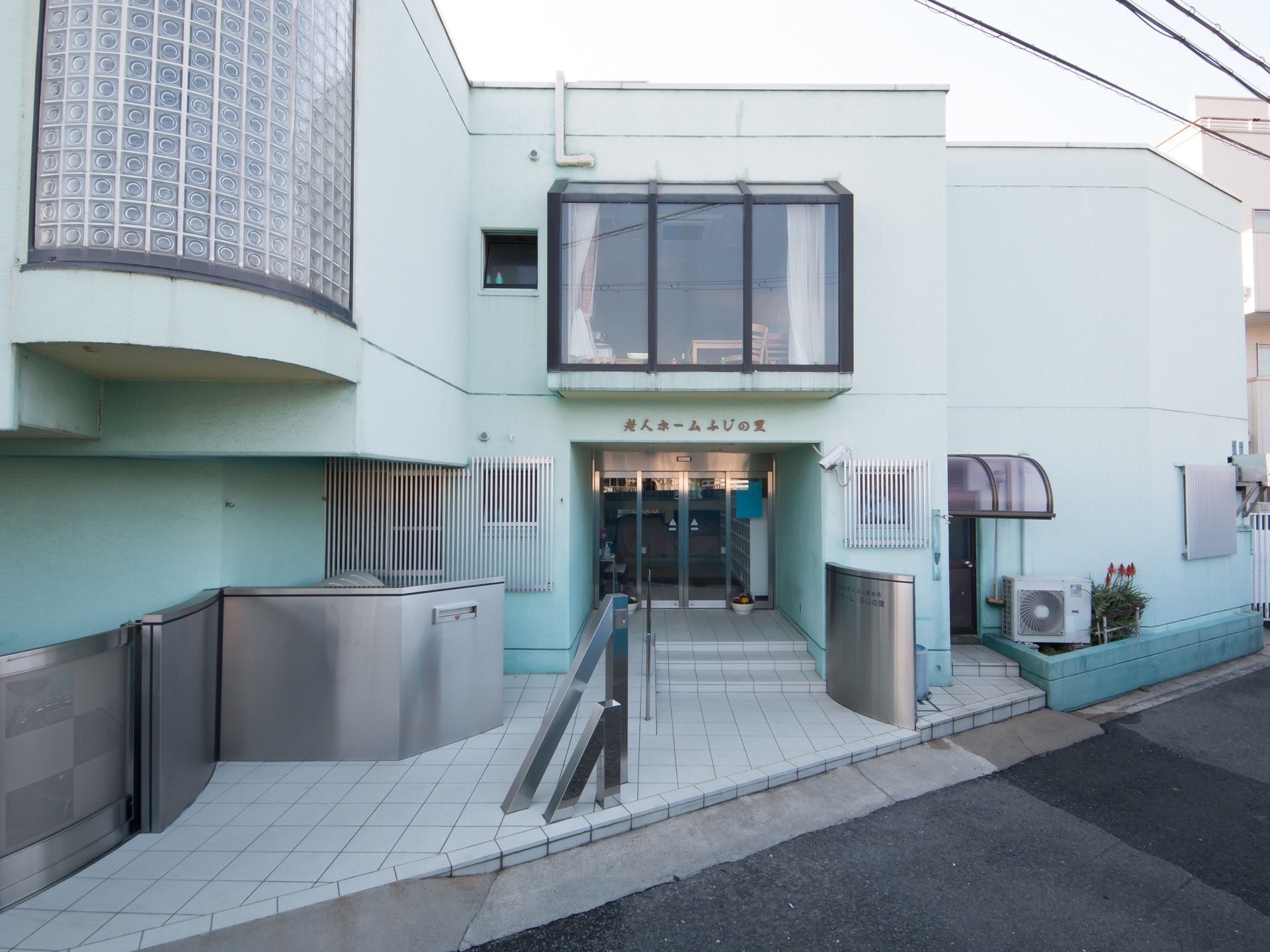 グリーンの２階建ての建物になっていて、玄関の上の壁に施設名が書かれている。１階の窓にはアルミの面格子が取りつけられている。