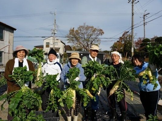 農作物を持って笑顔で高齢者が並んでいる写真。施設内の外スペース