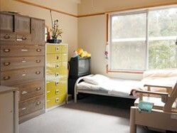 寝具付きのベッドが窓際に置かれ、黄色いタンスや木製の大きなタンスがあり、ポータブルトイレも設置されている。ベッドの上の方にはテレビもある。