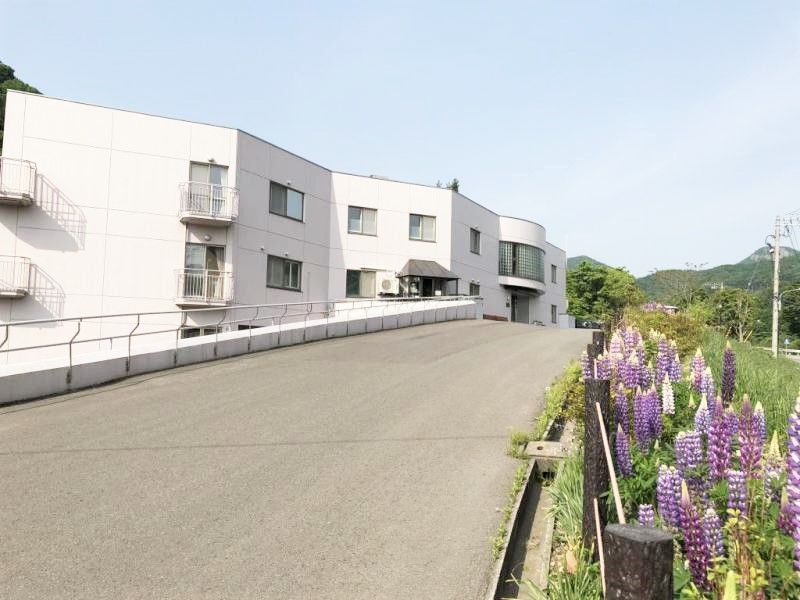 2階建ての建物へと続く道路の右側には紫の花が咲き、山々も見える。