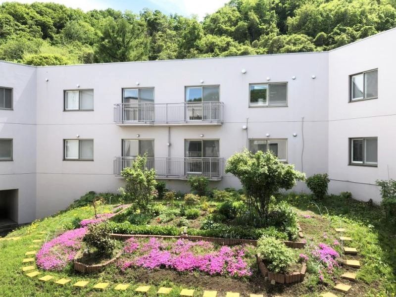 中庭には紫の花や木が植えられ、周囲をレンガブロックで囲っている。2階建ての建物の後ろには山も見える。