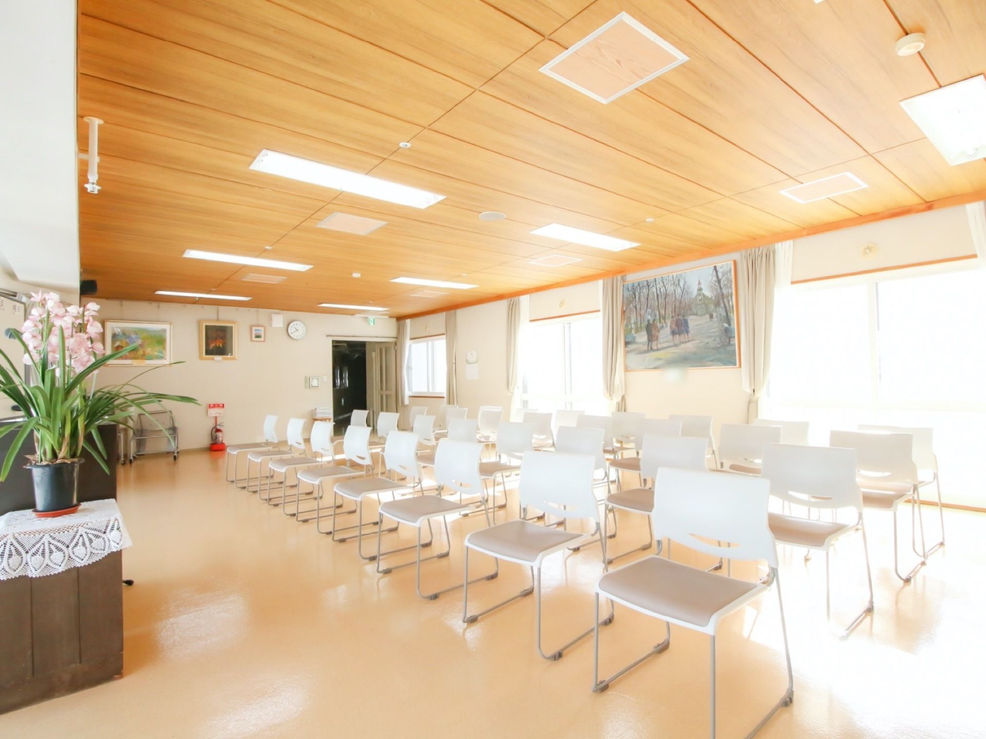 広い室内に、白い椅子がたくさん並べられている。壁には額絵が飾られており、消火器や蘭の鉢植えが置かれている。