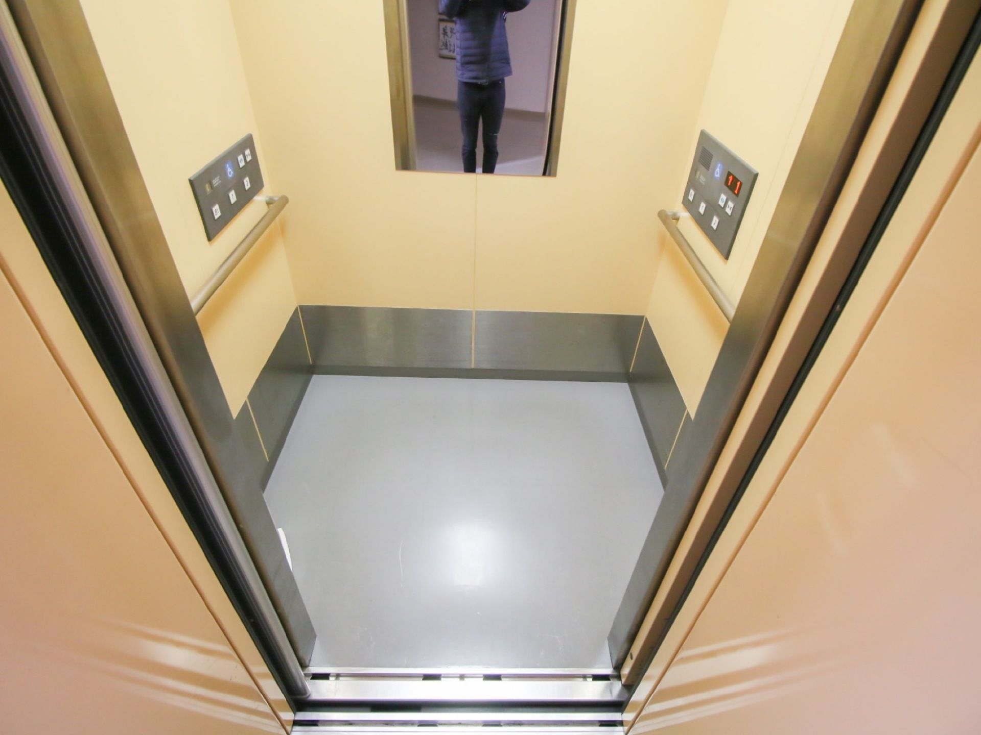 ベージュ色の壁とグレーの床のエレベーターで、奥の壁に縦長の鏡が備わっている。廊脇の壁には操作パネルと、銀色の手すりが設置されている。