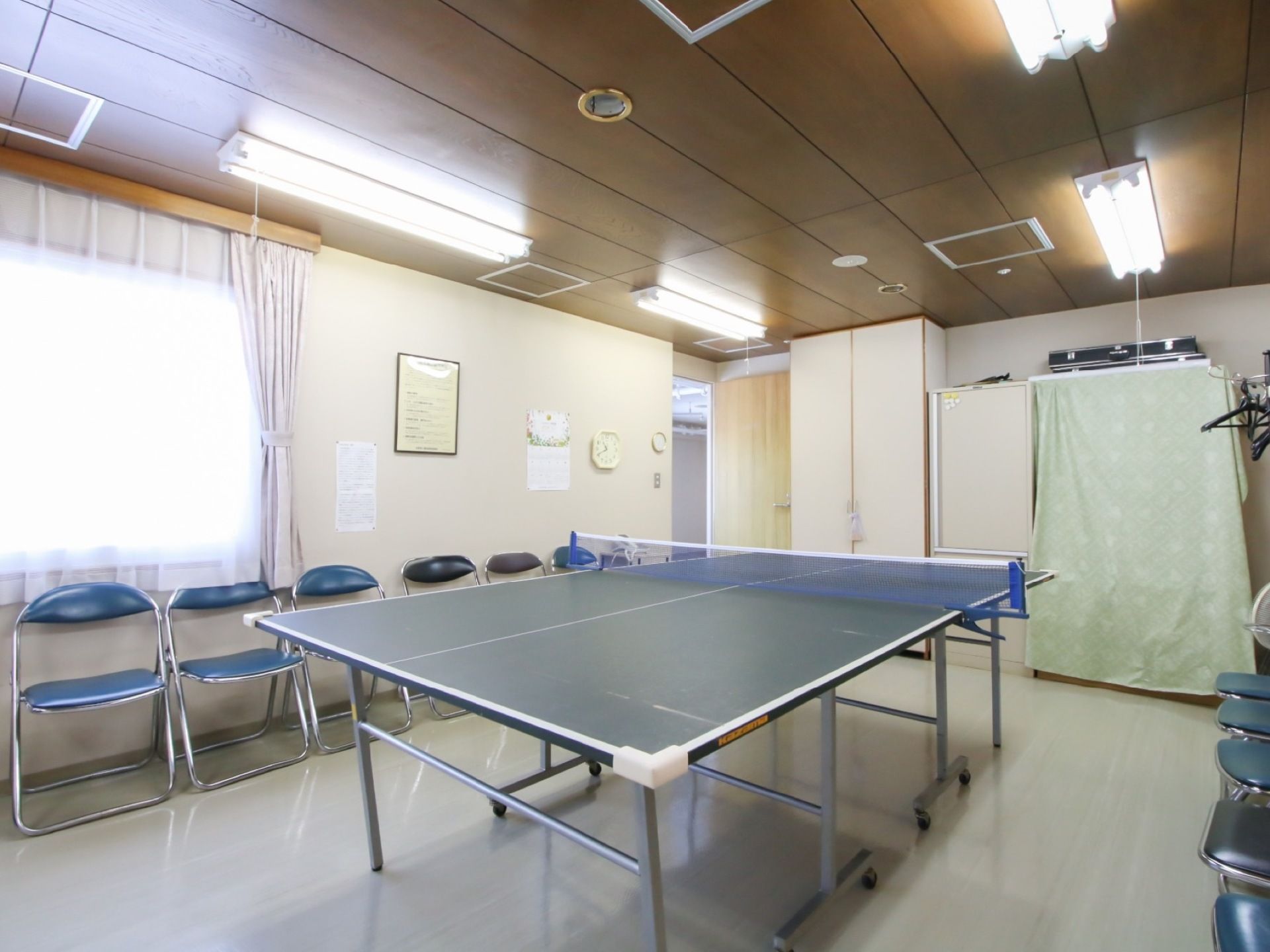 部屋の中央に卓球台があり、両脇の壁際に椅子が並んでいる。奥の壁の前にはタンスやハンガーラックなどが置かれている。