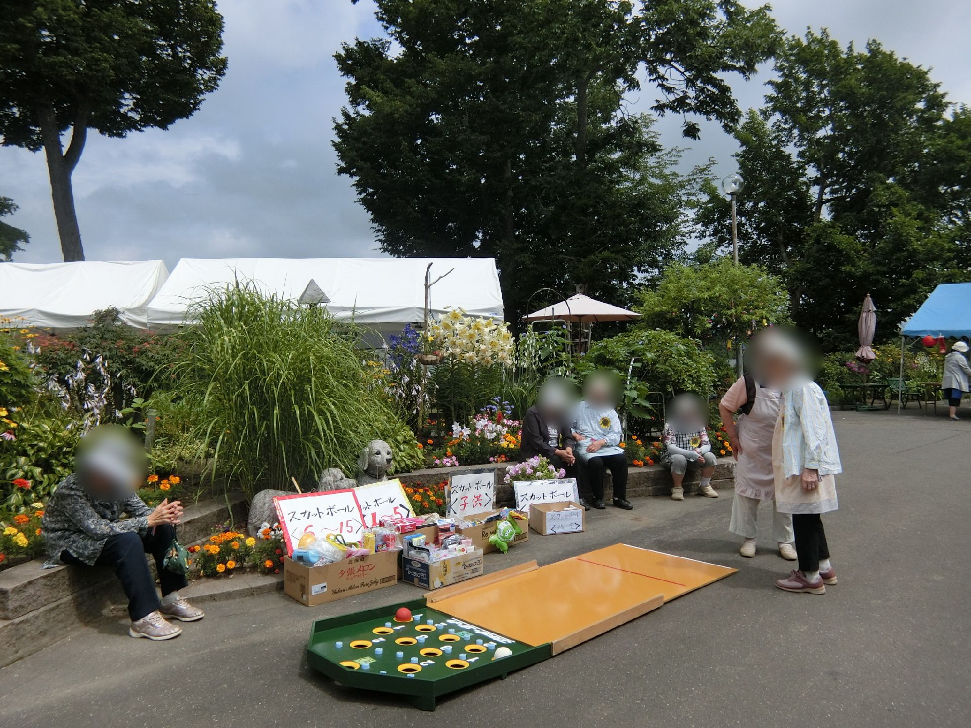 帽子をかぶった高齢者があちこちにいる。屋外の道路のような場所には夜店のようなゲームをする場所があり、奥には白いテントが立っている。