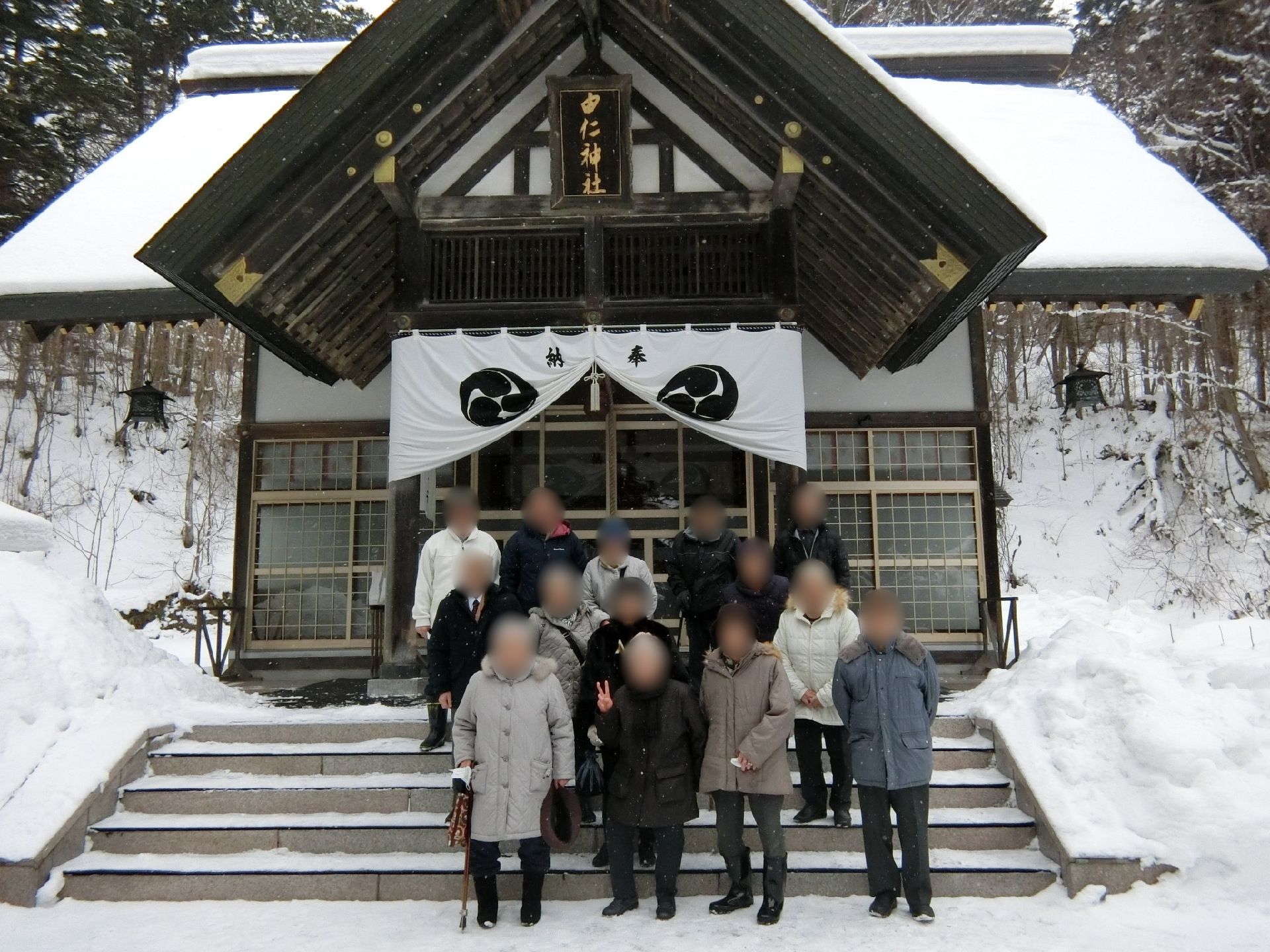 雪の積もった境内で社をバックに記念撮影に応じている高齢者たち。厚手のジャケットを着たり、帽子をかぶったりと、寒さ対策も万全。