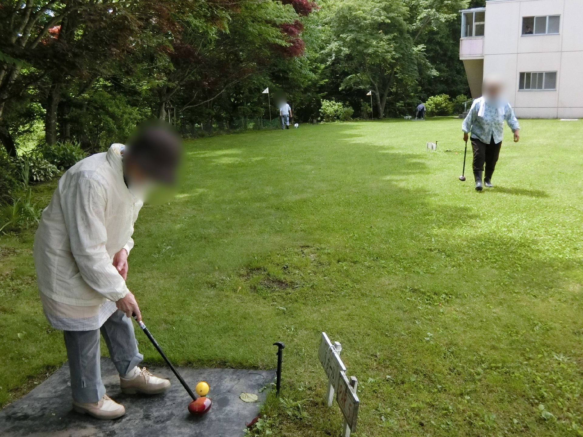 帽子をかぶって長袖長ズボンを身につけた高齢女性が、芝生が敷かれた広いスペースでパークゴルフを楽しんでいる様子。奥から一人こちらに歩いてきている。