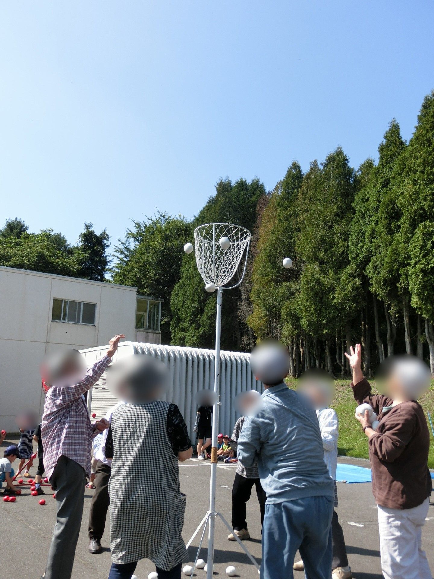 敷地は大きな緑の木に囲まれている。白い建物のそばで玉入れが行われていて、たくさんの高齢者が一生懸命球を投げている様子。