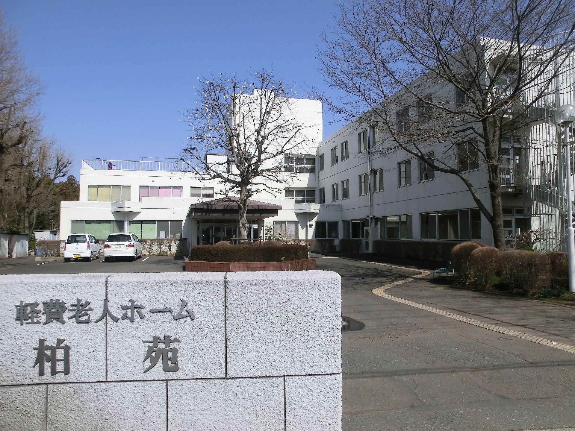 駐車場付きの建物。白を基調とした３階建ての建物の玄関には車寄せがあり、建物の周りには木が植えられている。