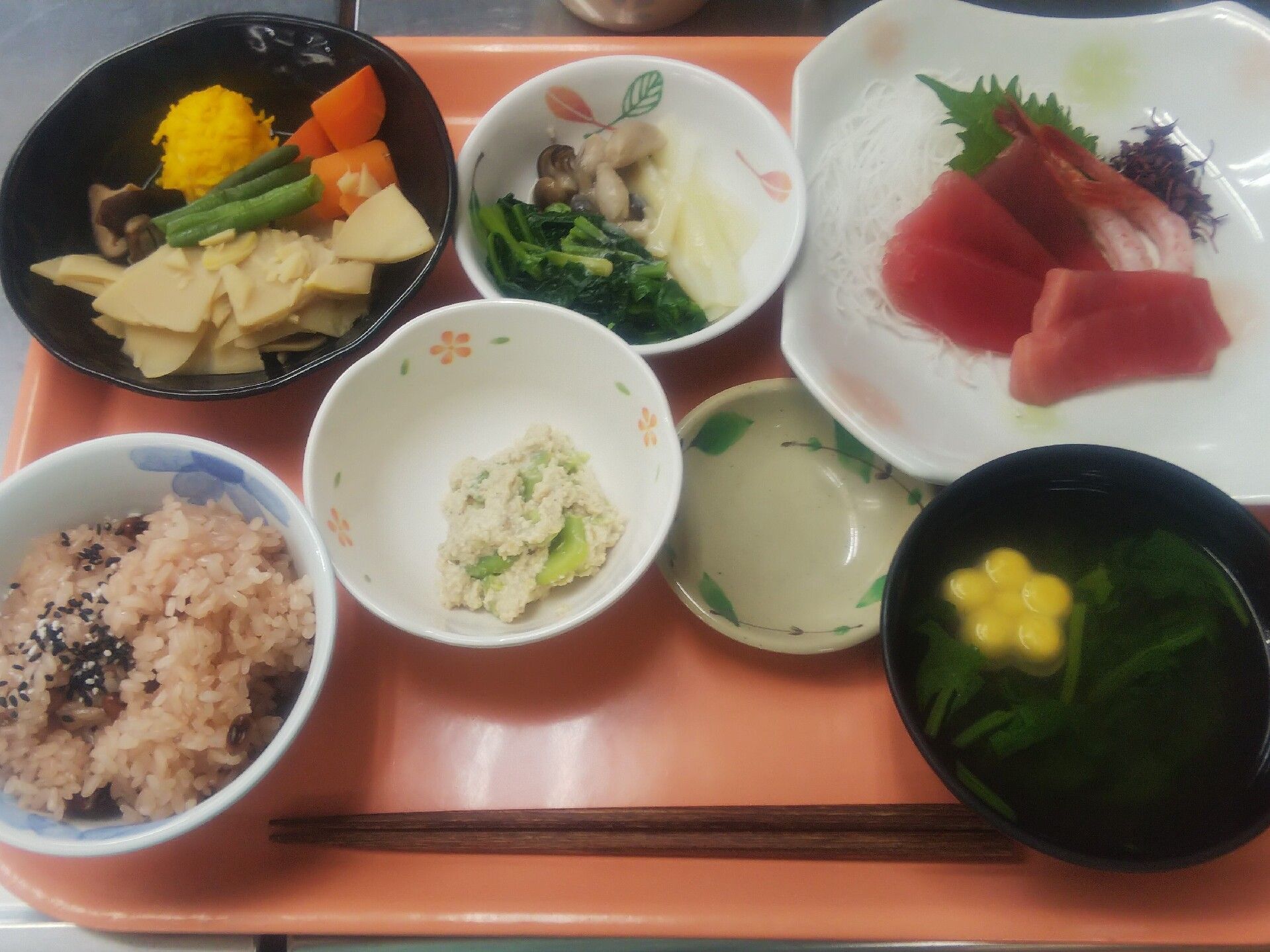 マグロの刺身や赤飯、吸い物、煮物、野菜の和え物などが並んで置かれているオレンジのトレー。