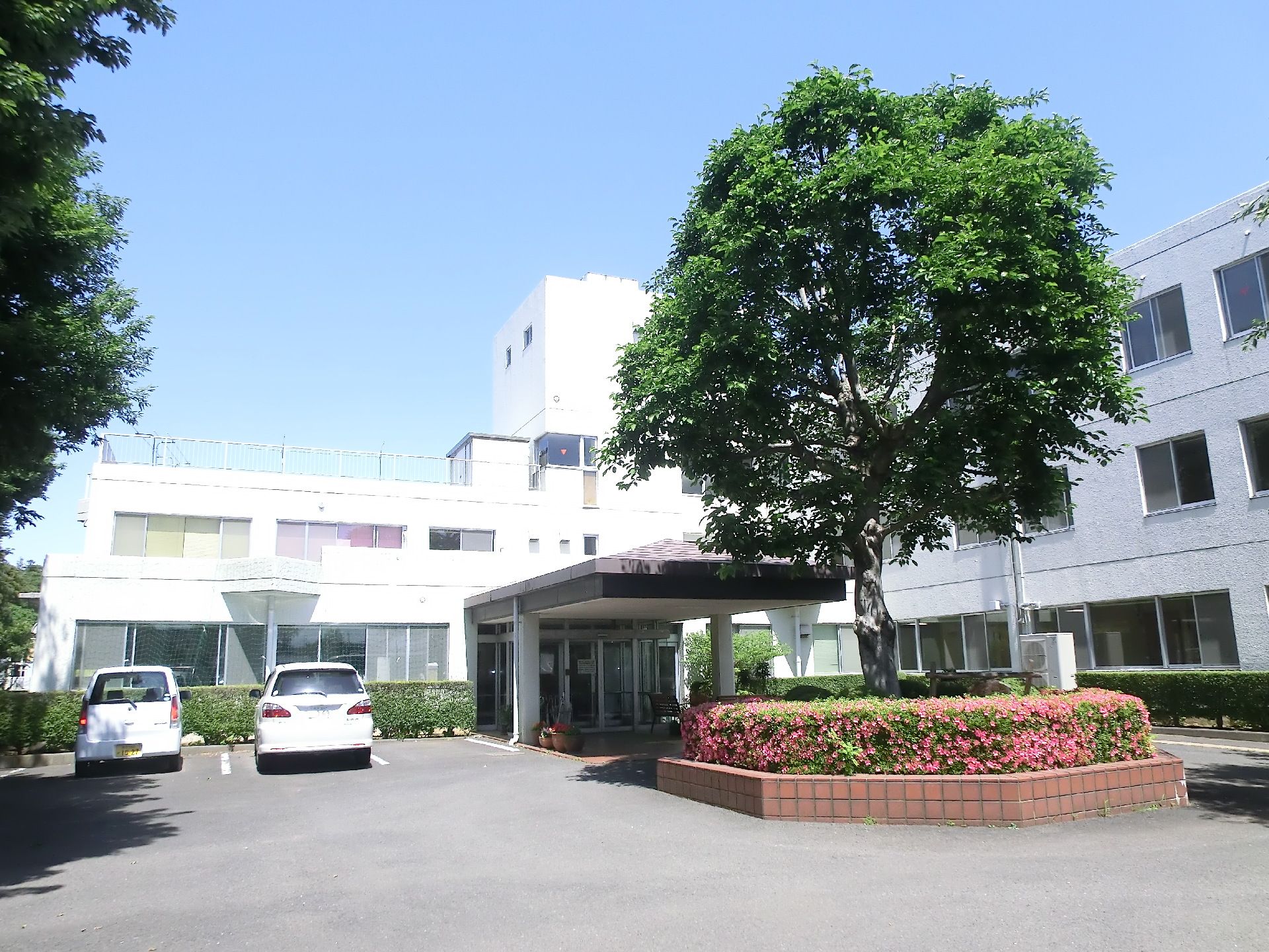 エル字型の白を基調とした３階建ての建物の前や周りには草木が植えられていて、駐車場もある。