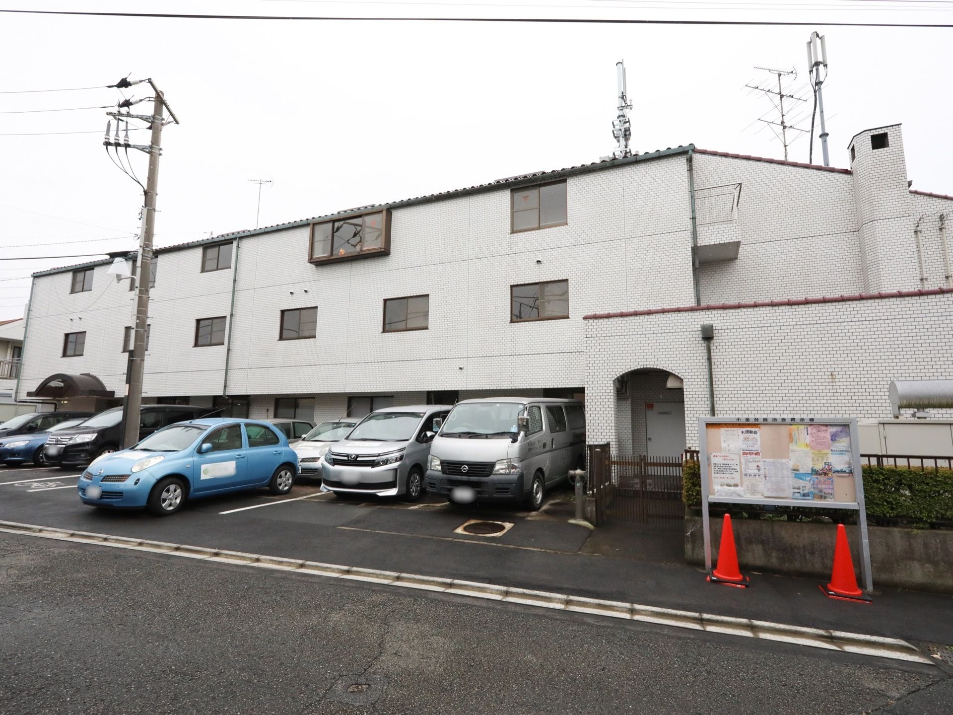 道路に面した、白い３階建ての建物である。正面に駐車スペースを設けている。横側にグレー枠の掲示版があり、さまざまな広告が貼られている。